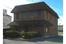 Two-story brick building with a dark roof, several narrow windows, and a small parking area in front. Bushes line the front of the building. Blue sky in the background.